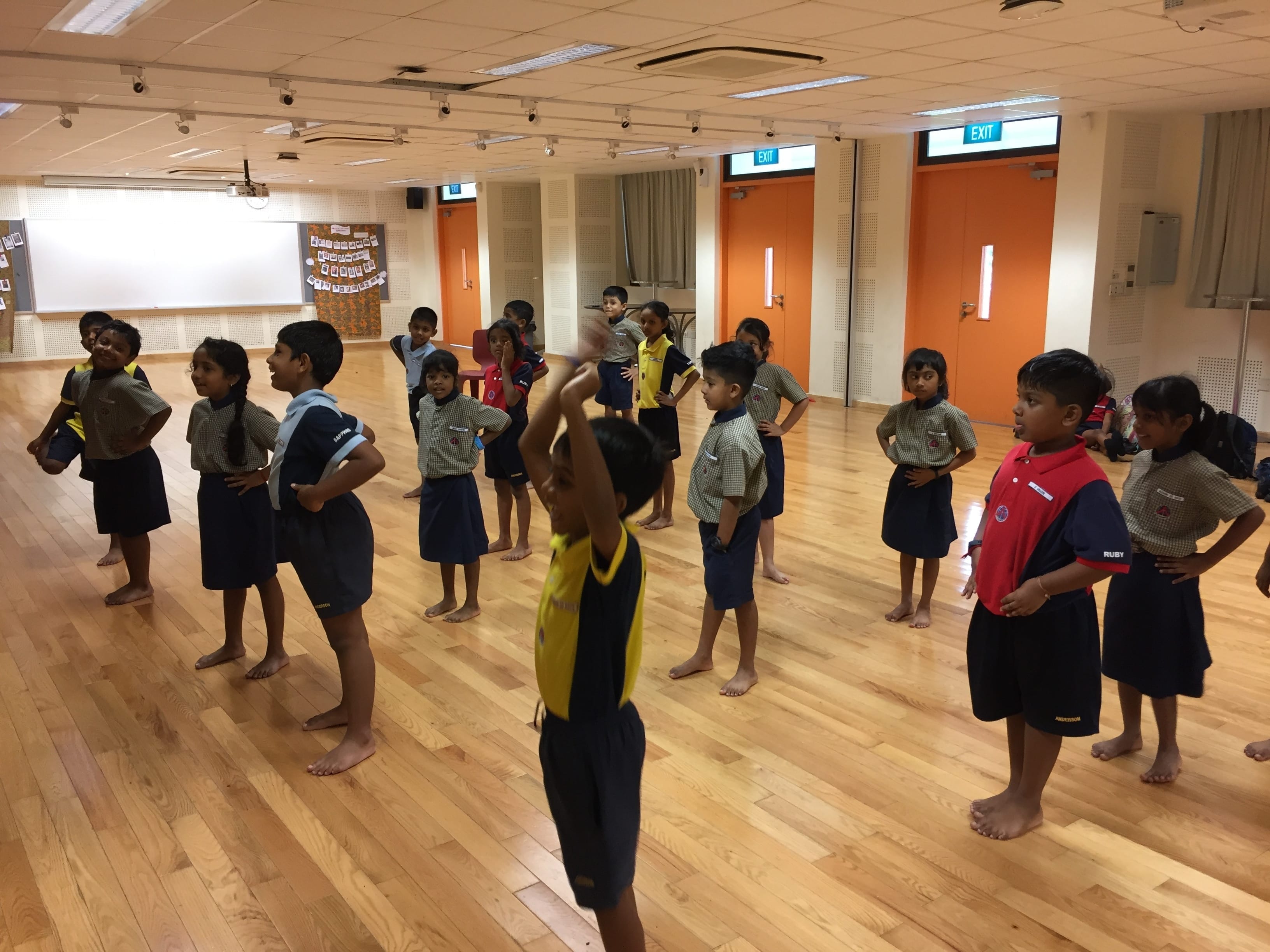 Young children in school uniforms stand barefoot on a wooden floor in a hall, one child raising arms overhead.